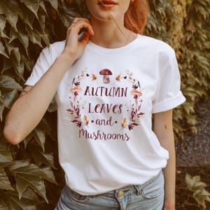 May include: A white short-sleeved t-shirt featuring a central graphic of autumn leaves and mushrooms arranged in a circular wreath. The text "AUTUMN LEAVES and MUSHROOMS" is printed in dark red lettering in the centre of the botanical design.