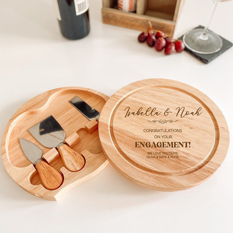 May include: A wooden cheese board with a round top and a separate compartment for cheese knives. The top of the board is engraved with the text "Isabella & Noah", "Congratulations on your Engagement!" and "We love you guys, Olivia & Kate & Alfie".