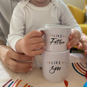 May include: Two white ceramic mugs with black text and gold stars. The top mug says "Like Father" and the bottom mug says "Like Son".