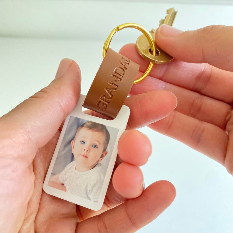 Puede incluir: Un llavero de cuero marr&oacute;n con la palabra "Granda" grabada en &eacute;l. El llavero tiene un peque&ntilde;o marco de fotos blanco con una foto de un beb&eacute; dentro. El llavero est&aacute; unido a un anillo de llave dorado.