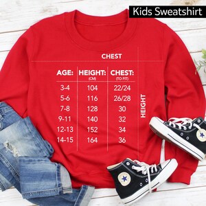 Op de afbeelding: Rood kindertrui met een maattabel op de voorkant. De tabel toont leeftijd, lengte in centimeters en borstomvang in inches. De tabel is getiteld "Kids Sweatshirt".