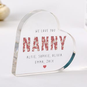Puede incluir: Un bloque acrílico transparente en forma de corazón. La palabra "NANNY" está en letras grandes con estampado floral. El texto "WE LOVE YOU" está arriba, con nombres y un corazón debajo.
