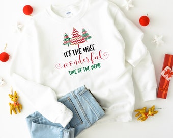 May include: A white sweatshirt with the text "It's the most wonderful time of the year" and a graphic of three Christmas trees. The sweatshirt is paired with blue jeans, red ornaments, and decorative stars, suggesting a festive holiday theme.