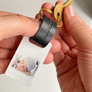 Puede incluir: Un llavero de cuero negro con la palabra "GRANDPA" grabada en &eacute;l. El llavero tiene un peque&ntilde;o portafotos rectangular blanco con una foto de un beb&eacute; durmiendo con un animal de peluche.