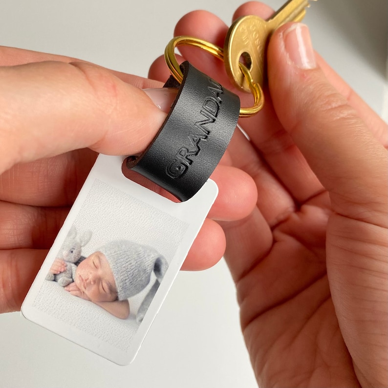 Puede incluir: Un llavero de cuero negro con la palabra "GRANDPA" grabada en &eacute;l. El llavero tiene un peque&ntilde;o portafotos rectangular blanco con una foto de un beb&eacute; durmiendo con un animal de peluche.