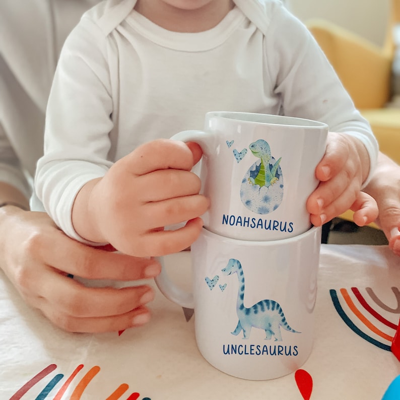 Pu&ograve; includere: Due tazze di ceramica bianca impilate. La tazza superiore presenta un dinosauro blu che esce da un uovo, con il nome "NOAHSAURUS" sotto. La tazza inferiore ha un dinosauro blu con il nome "UNCLESAURUS".