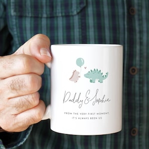 May include: White ceramic mug featuring two cartoon dinosaurs, one holding a balloon. The mug has the text "Daddy & Sophie" and the phrase "From the very first moment, it's always been us."