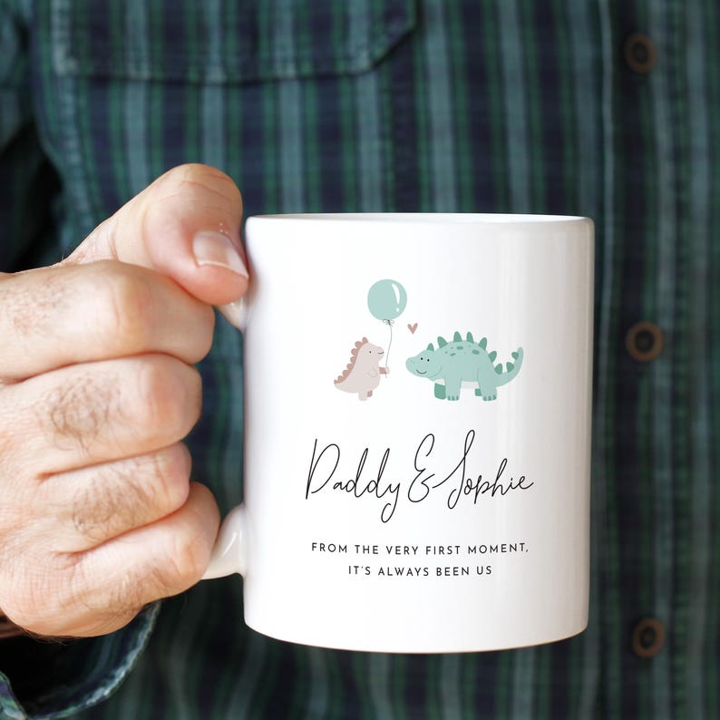 May include: White ceramic mug featuring two cartoon dinosaurs, one holding a balloon. The mug has the text "Daddy & Sophie" and the phrase "From the very first moment, it's always been us."