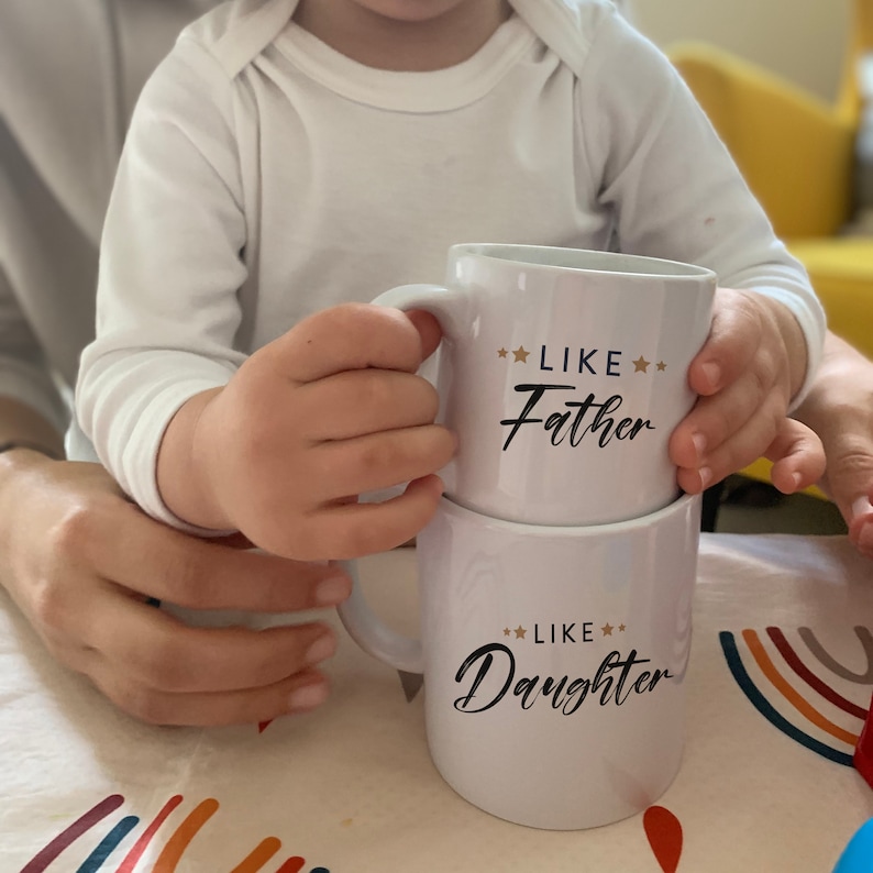 May include: Two white ceramic mugs stacked on top of each other. The top mug has the text "Like Father" with two gold stars on either side of the word "Like". The bottom mug has the text "Like Daughter" with two gold stars on either side of the word "Like".
