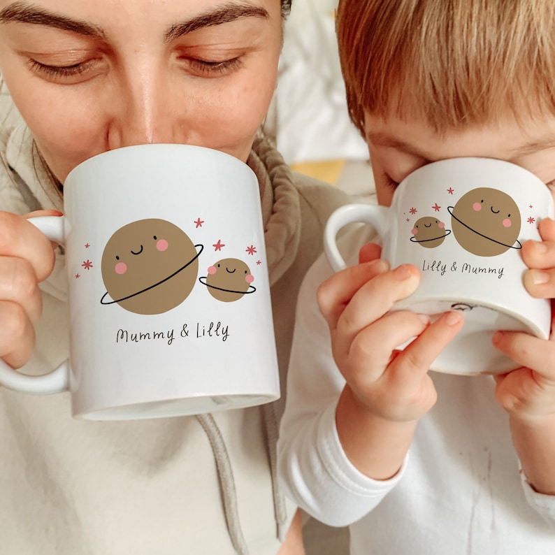 May include: Two white ceramic mugs with a planet design. The larger mug says "Mummy & Lilly" and the smaller mug says "Lilly & Mummy".