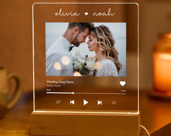 Puede incluir: Un marco de fotos iluminado de acrílico transparente con una foto de boda de una pareja, una interfaz de reproductor de música y el texto "olivia ♥ noah" en la parte superior. El marco tiene una base de madera.