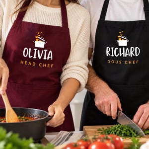 Può includere: Un grembiule bordeaux con la scritta OLIVIA HEAD CHEF e un grafico di una pentola bianca con cuori arancioni, e un grembiule nero con la scritta RICHARD SOUS CHEF e un grafico di una pentola bianca con cuori arancioni, entrambi con lacci.
