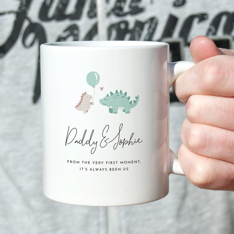 May include: White ceramic mug featuring a dinosaur theme. The mug has a cartoon illustration of two dinosaurs, a pink one holding a balloon and a green one. The text "Daddy & Sophie" is written in cursive, with additional text below.