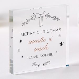 Puede incluir: Un bloque acrílico transparente con las palabras "MERRY CHRISTMAS tía & tío LOVE SOPHIE" en texto negro y color cobre. El bloque presenta un lazo y un diseño floral en la parte superior y acentos de estrellas.
