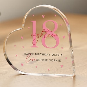 May include: A clear heart-shaped acrylic paperweight with the number 18 in pink and the word "eighteen" in pink script. The text "Happy Birthday Olivia Love Auntie Sophie" is printed below the number. The paperweight is decorated with small pink hearts.