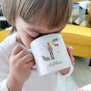Puede incluir: Taza de cerámica blanca con una ilustración colorida de un cascanueces, adornos navideños y vegetación. La taza está personalizada con el nombre "Arthur" escrito en cursiva.