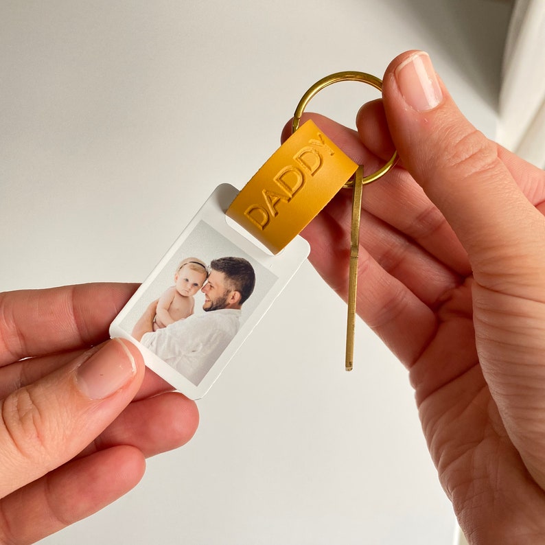 K&ouml;nnte beinhalten: Ein personalisierter Schl&uuml;sselanh&auml;nger mit einem Foto von Vater und Kind. Der Schl&uuml;sselanh&auml;nger hat ein senffarbenes Lederetikett mit dem gepr&auml;gten Wort "DADDY". Das Foto ist rechteckig und an einem Messing-Schl&uuml;sselring befestigt.