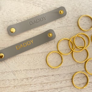 K&ouml;nnte beinhalten: Zwei graue Leder-Schl&uuml;sselanh&auml;nger mit dem Wort "DADDY" in Gold gepr&auml;gt. Jeder Schl&uuml;sselanh&auml;nger hat einen goldenen Druckknopfverschluss. Mehrere goldfarbene Schl&uuml;sselringe liegen daneben, was auf ein Set hindeutet.
