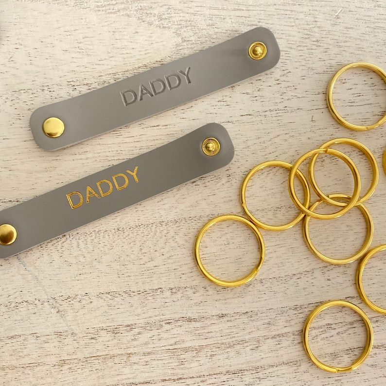 K&ouml;nnte beinhalten: Zwei graue Leder-Schl&uuml;sselanh&auml;nger mit dem Wort "DADDY" in Gold gepr&auml;gt. Jeder Schl&uuml;sselanh&auml;nger hat einen goldenen Druckknopfverschluss. Mehrere goldfarbene Schl&uuml;sselringe liegen daneben, was auf ein Set hindeutet.