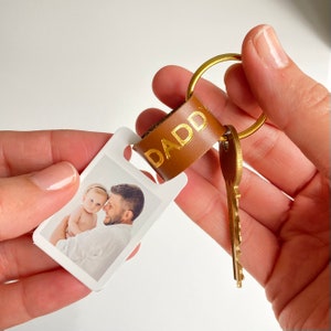 May include: A brown leather keychain with a gold metal key ring and a photo card. The photo card features a father and daughter. The leather strap has the word "DADDY" embossed in gold lettering.