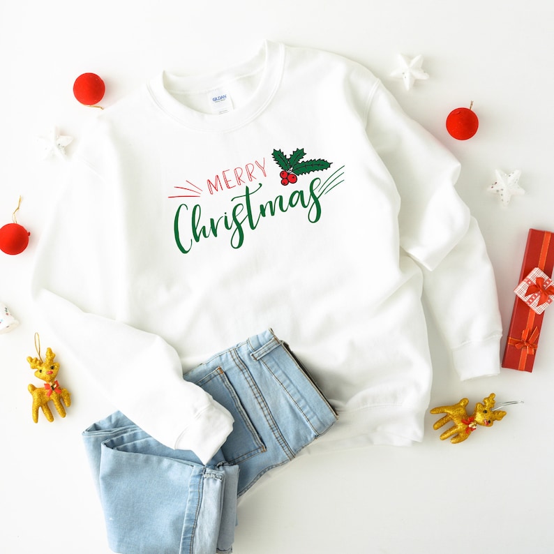 Op de afbeelding: Witte trui met een groene hulsttak en rode bessen met de tekst "Merry Christmas" in groene en rode letters.