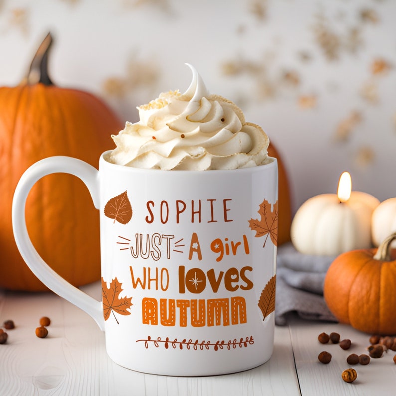 May include: White ceramic mug with a brown handle. The mug has a design of orange and brown leaves and the text "Sophie Just A Girl Who Loves Autumn". The mug is filled with whipped cream.