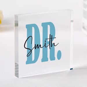 Può includere: Un blocco di acrilico trasparente con il testo "DR. Smith" in scrittura corsiva nera e "DR" in grandi lettere blu.