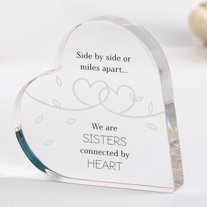 Puede incluir: Un pisapapeles de acrílico transparente en forma de corazón con el texto "Lado a lado o a kilómetros de distancia... Somos HERMANAS conectadas por el CORAZÓN" y un diseño de dos corazones entrelazados con hojas.