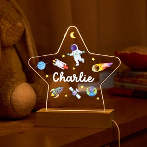 May include: A clear acrylic star-shaped night light with a wooden base. The star has a space theme with an astronaut, rockets, planets, and stars. The name "Charlie" is written on the star.