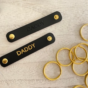 K&ouml;nnte beinhalten: Zwei schwarze Leder-Schl&uuml;sselanh&auml;nger mit dem Wort "DADDY" gepr&auml;gt und goldfarbener Hardware. Die Schl&uuml;sselanh&auml;nger sind rechteckig mit abgerundeten Enden und Druckkn&ouml;pfen. Mehrere goldfarbene Schl&uuml;sselringe sind ebenfalls sichtbar.