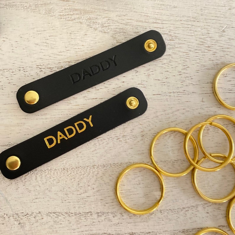 K&ouml;nnte beinhalten: Zwei schwarze Leder-Schl&uuml;sselanh&auml;nger mit dem Wort "DADDY" gepr&auml;gt und goldfarbener Hardware. Die Schl&uuml;sselanh&auml;nger sind rechteckig mit abgerundeten Enden und Druckkn&ouml;pfen. Mehrere goldfarbene Schl&uuml;sselringe sind ebenfalls sichtbar.