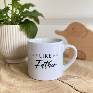 May include: White ceramic mug with a black and gold design that says "Like Father" with stars on either side of the word "Like".