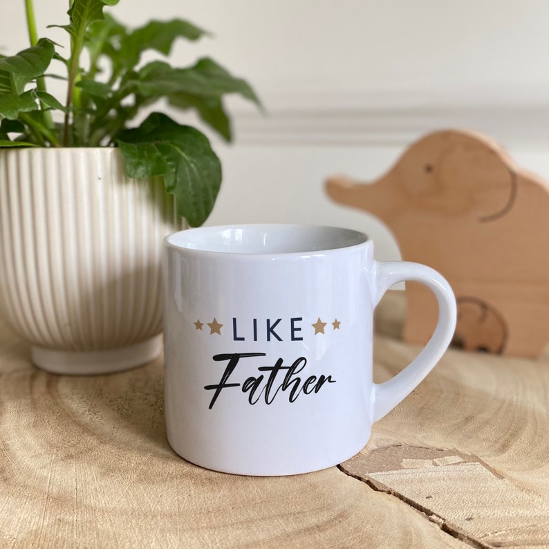 May include: White ceramic mug with a black and gold design that says "Like Father" with stars on either side of the word "Like".
