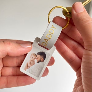 Puede incluir: Llavero de cuero blanco con una inscripci&oacute;n 'DADDY' dorada y una foto de un padre sosteniendo a un beb&eacute;.