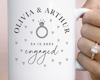 Op de afbeelding: Witte keramische mok met de namen "OLIVIA & ARTHUR" en het woord "engaged" in zwart schrift. Een diamantringafbeelding en de datum 24.12.2025 zijn ook te zien. Kleine hartafbeeldingen omringen de tekst.