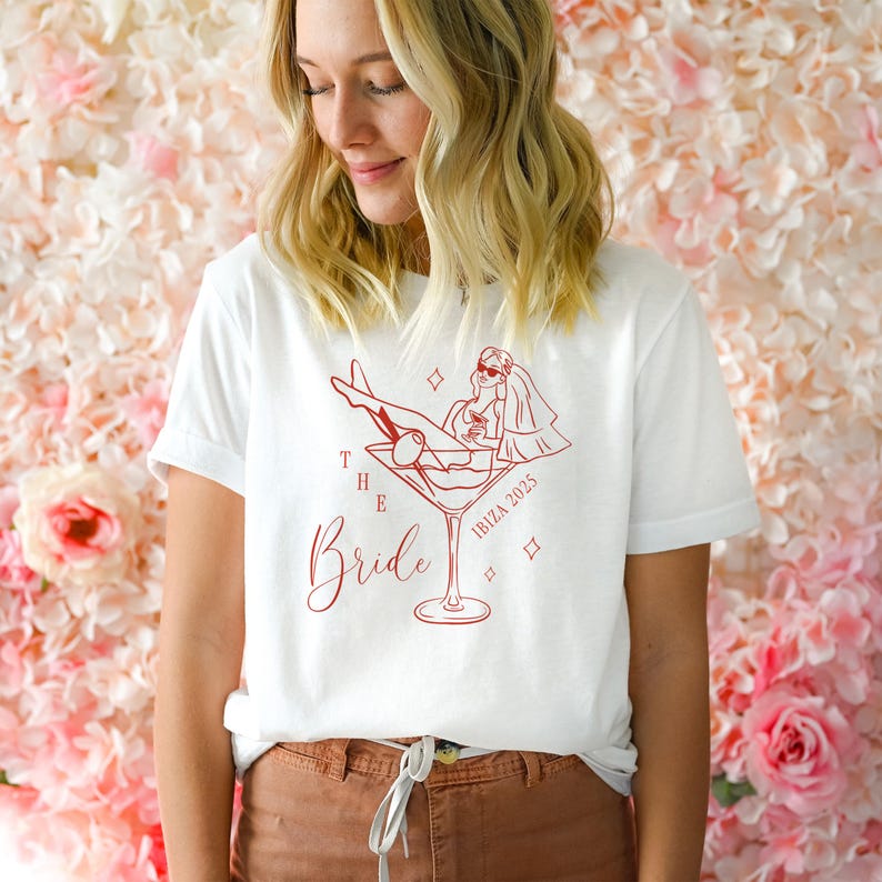 May include: White t-shirt with a red graphic of a bride in a martini glass, wearing sunglasses and a veil. The text "The Bride" is written in cursive, with "Ibiza 2025" below the graphic. The shirt has short sleeves.