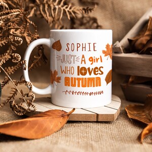 May include: White ceramic mug with a handle. The mug has a design of orange and brown autumn leaves and the text "Sophie Just A Girl Who Loves Autumn" in orange and brown.