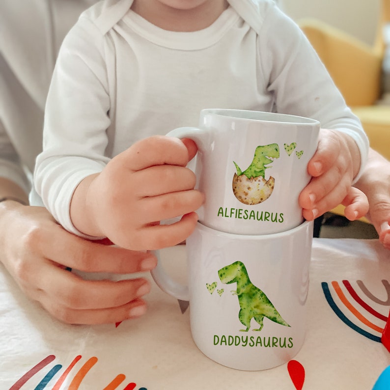 Pu&ograve; includere: Due tazze di ceramica bianca impilate. La tazza superiore presenta un dinosauro verde che esce da un uovo, con la scritta "ALFIESAURUS". La tazza inferiore ha un dinosauro verde con la scritta "DADDYSAURUS".