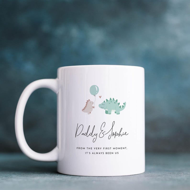 May include: White ceramic mug with a handle, featuring a whimsical design of two dinosaurs, a balloon, and the text "Daddy & Sophie". Below the names, the text reads "FROM THE VERY FIRST MOMENT, IT'S ALWAYS BEEN US."