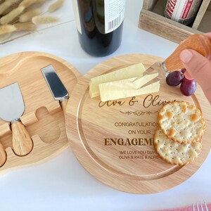 May include: A wooden cheese board with a heart-shaped cutout and a smaller round board with a knife, cheese, crackers, and grapes. The round board has the text "Ava & Olivia Congratulations on your Engagement We love you guys Olivia & Kate & Alfie" engraved on it.