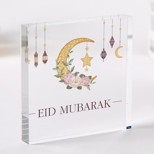 May include: A white square acrylic block with a gold crescent moon and star design. The moon is decorated with floral accents and the text "EID MUBARAK" is printed in brown below the design.