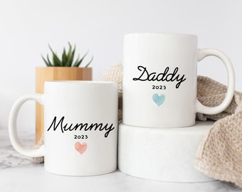 May include: Two white ceramic mugs with black calligraphy. The mug on the left says "Mummy 2023" with a pink watercolor heart. The mug on the right says "Daddy 2023" with a blue watercolor heart.