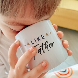 May include: A white ceramic mug with the text "Like Father" in black cursive lettering and two gold stars on either side of the word "Like".