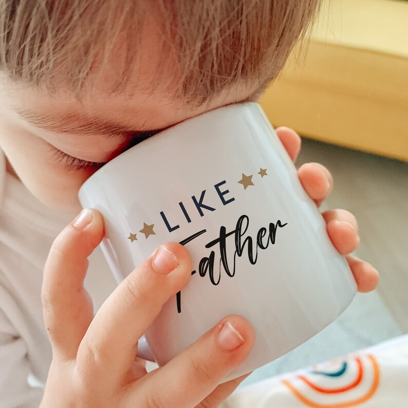 May include: A white ceramic mug with the text "Like Father" in black cursive lettering and two gold stars on either side of the word "Like".