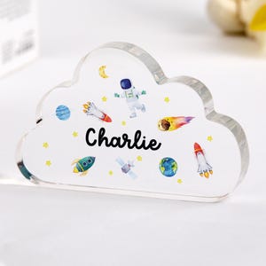 Könnte beinhalten: Ein wolkenförmiger Acrylblock mit Weltraum-Design. Das Design umfasst Raketen, Planeten, Sterne und einen Astronauten. Der Name "Charlie" steht in schwarzer Schrift über der Mitte des Blocks.