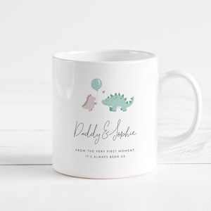 May include: White ceramic mug featuring a dinosaur theme. The mug has a light blue balloon and two dinosaurs, one pink and one green. The text "Daddy & Sophie" and a quote are printed on the mug.