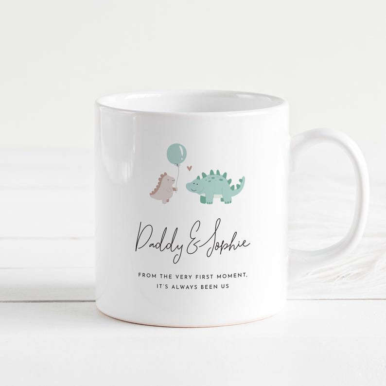 May include: White ceramic mug featuring a dinosaur theme. The mug has a light blue balloon and two dinosaurs, one pink and one green. The text "Daddy & Sophie" and a quote are printed on the mug.
