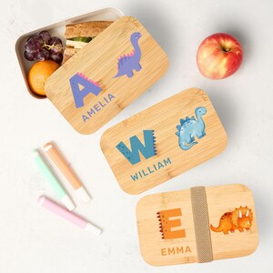 May include: Three bamboo lunch boxes with dinosaur designs. One box has the name "Amelia" with a purple dinosaur, another says "William" with a blue dinosaur, and the third says "Emma" with an orange dinosaur. The boxes are on a white surface with fruit and markers.