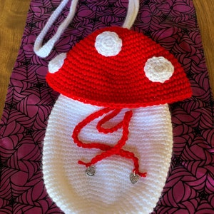 Może przedstawiać: Worek w kształcie grzyba wykonany na szydełku, z białym korpusem i czerwoną górą. Worek ma biały sznurek do ściągania i dwa srebrne, sercowate breloczki.