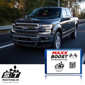 May include: A black Ford F-150 pickup truck driving on a road. The truck has a black grille with a chrome Ford logo. The truck is driving on a road with trees on either side. The truck has a black and blue performance chip with the text "MAXX BOOST Performance Chip" and a QR code. The chip is from SawyerTuning.com.